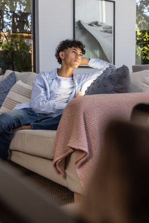 Man sitting on beige sofa with pink throw, gazing through large glass window at greenery. Contemplation, relaxation, interior design, cozy, serenity, contemporary, lifestyleの写真素材