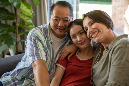 Mature parents with adult daughter sitting on dark couch by potted plant in living room, smiling. Family, togetherness, warmth, harmony, trio, daylight, windowの写真素材