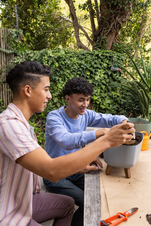 Male friends filling gray pot and orange planter with potting soil at wooden backyard table....
