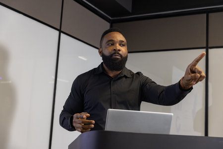African American man pointing forward while presenting with silver laptop at podium in meeting room....