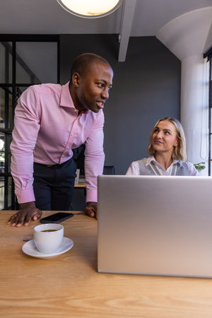Diverse coworkers discussing project details while leaning on desk with silver laptop in office....