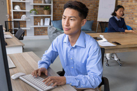 Asian coworkers typing on keyboard at open-plan office desk, light-blue shirt visible....