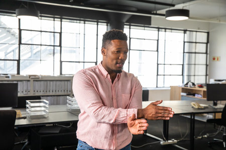 Adult African American man wearing pink shirt and jeans, standing and gesturing at office monitors....