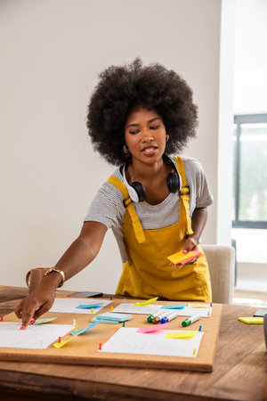 African American woman wearing yellow overalls at home desk sorting sticky notes on corkboard....