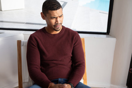 Man sitting pensively on woven-seat chair in living room by large window overlooking pool deck. Contemplation, relaxation, home, interior, modern, lifestyle, comfortの写真素材