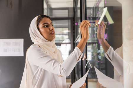 Indian woman in headscarf writing on glass partition with neon marker, holding paper in office. Collaboration, brainstorming, innovation, workspace, professional, modern, creativityの写真素材