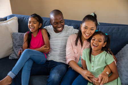 Diverse family of four laughing together on deep blue sofa in living room holding remote control....