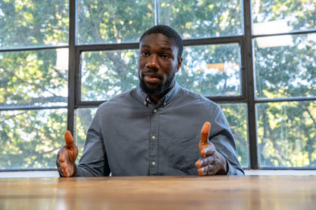 African American man gesturing while sitting at conference table by grid windows with sunlit trees....