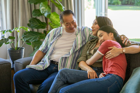 Diverse family sitting on gray sofa at home with plants parents holding daughter wearing wristwatch....