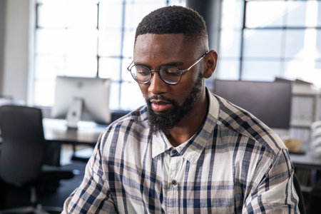 African American man wearing round glasses, plaid shirt working at desk in office with monitors. Workspace, screens, daylight, productivity, focus, contemporary, corporateの写真素材