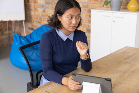 Asian woman sitting at coworking in navy sweater holding tablet on small stand on table gesturing....