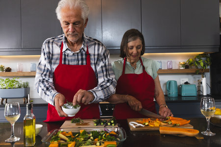 Senior couple cooking at kitchen island wearing red aprons, sprinkling herbs and slicing carrots....