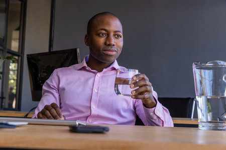 African American adult man sitting at office table holding smartphone and water glass, copy space. Professional, workspace, modern, technology, corporate, productivity, businessの写真素材