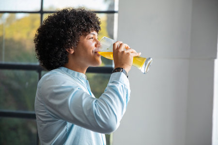 Adult African American man in light blue shirt standing by window drinking orange juice from glass. Modern, lifestyle, minimalism, wellness, refreshment, natural, serenityの写真素材