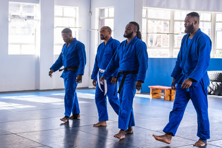 African American male martial artists walking barefoot across mat in blue gis with belts at studio....
