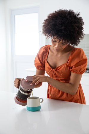African American woman pouring coffee from French press into two-toned mug at kitchen counter. Modern, minimalist, lifestyle, interior, cozy, bright, domesticの写真素材