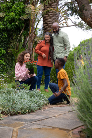 Diverse family kneeling and standing in lush backyard garden holding potted orange plant. Family,...