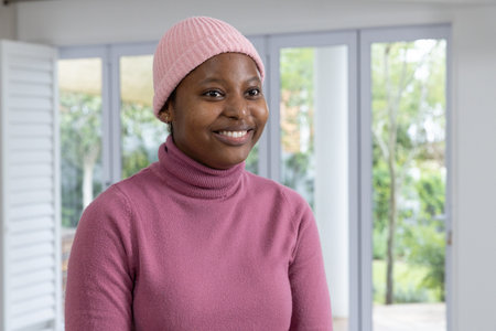 African American woman standing in bright room by glass doors wearing pink beanie and turtleneck....