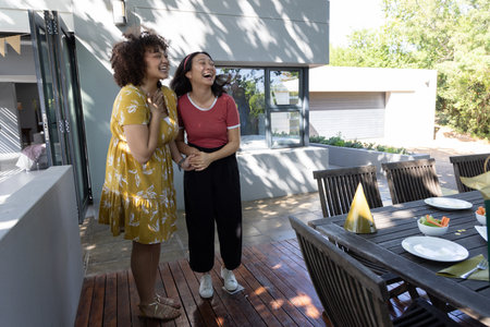 African American and Asian female friends laughing and holding hands on deck with dining table....