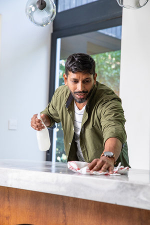 South Asian man wearing green shirt wiping marble countertop in kitchen with spray bottle and cloth....