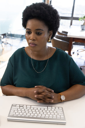 African American woman sitting at office desk in green blouse typing on wireless keyboard....