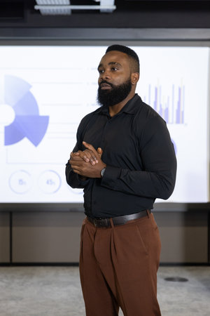 African American man wearing black shirt presenting charts on projection screen in meeting room....