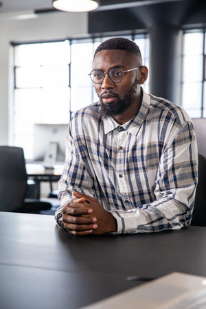 African American man sitting at conference table in modern office, wearing glasses and plaid shirt....