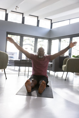 African American senior man stretching on yoga mat in maroon shirt in living room with windows....