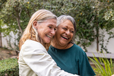 Diverse senior female friends hugging each other in residential garden with lush greenery backdrop....