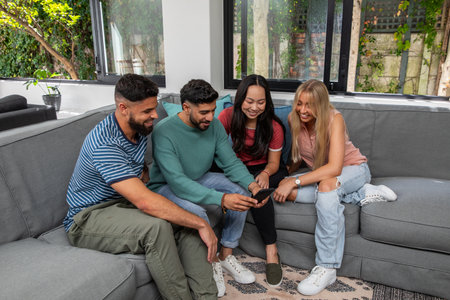 Diverse friends leaning in, sharing smartphone while sitting on gray sectional sofa in living room....