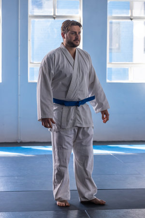 Male karate student standing barefoot wearing white gi blue belt in dojo on mats near windows....