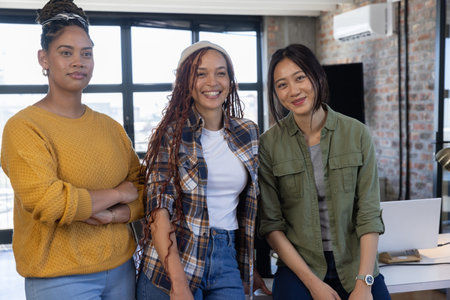 Diverse female coworkers standing in modern office overlooking cityscape with laptop and monitor....
