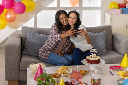 Diverse mother and teenage daughter taking selfie at home with smartphone and birthday cake....