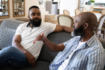 African American male friends chatting casually on gray sofa in living room with dining table....