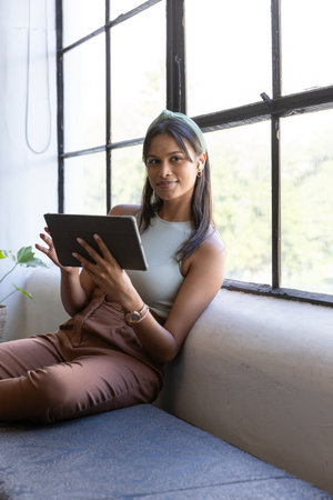 Asian woman holding tablet while sitting on bench by window in loft with earbuds and plant. Modern,...