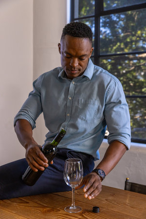 African American man pouring red wine from bottle into wine glass on wooden table at home. Casual,...