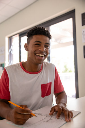 African American teenage boy writing in spiral-bound notebook with pencil at desk in classroom....