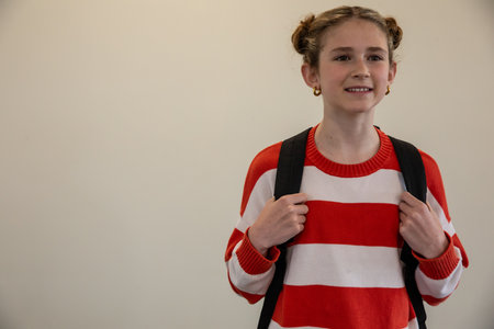 Child female student standing in school hallway holding black backpack straps, copy space. Youth,...