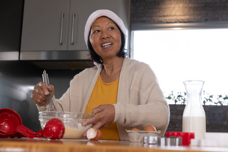 Senior Asian woman whisking batter at kitchen counter with glass bowl, red spoons, egg carton....
