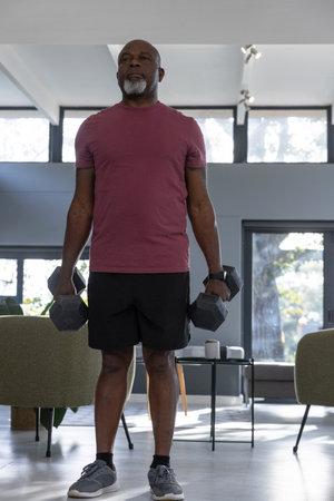 Senior African American man wearing athletic attire holding dumbbells at sides in living room gym. Fitness, strength, wellness, homegym, healthyaging, activeliving, vitalityの写真素材