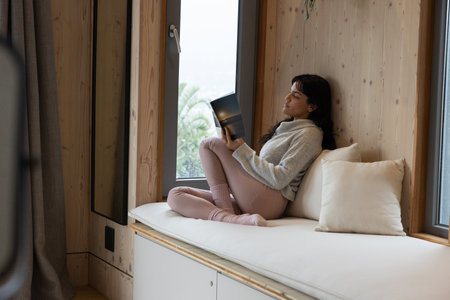 Hispanic woman sitting on bench with cushions reading book in reading nook by window, copy space. Cozy, serenity, sanctuary, relaxation, natural light, minimalist, wellnessの写真素材