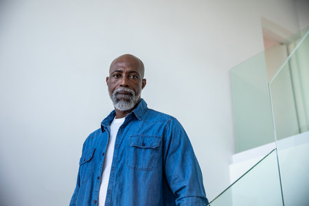 African American senior man standing by glass railing at staircase wearing denim shirt over T-shirt....
