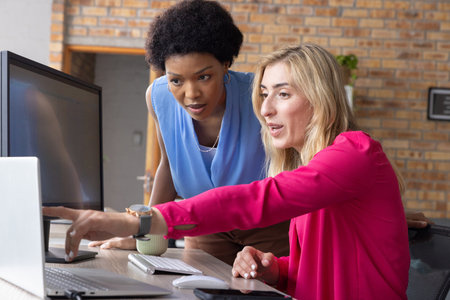 Diverse female coworkers collaborating at office desk with laptop and external monitor....