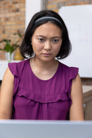 Asian woman working on laptop at office desk with brick accent wall, whiteboard, and potted plant....