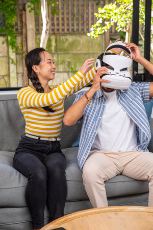 Indian and Chinese couple placing VR headset on head while sitting on grey sofa on patio....