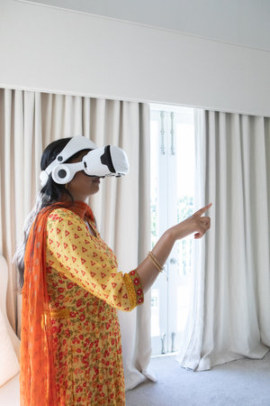 Indian woman wearing VR headset and traditional kurta reaching forward in living room, copy space....