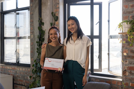 Diverse female colleagues in business attire standing in office holding framed certificate....