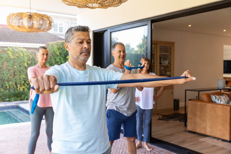 Senior friends holding blue resistance bands and stretching together in sportswear on patio by pool....