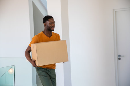 African American man carrying large cardboard box inside modern hallway with glass railing. Urban,...