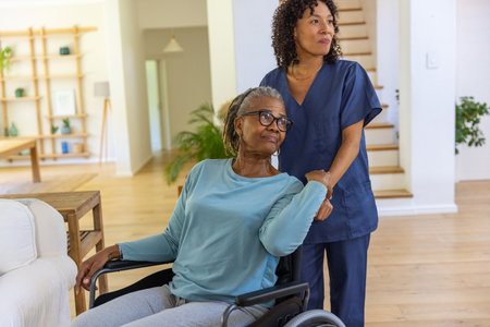 African American caregiver in scrubs assisting senior patient sitting in wheelchair in living room....